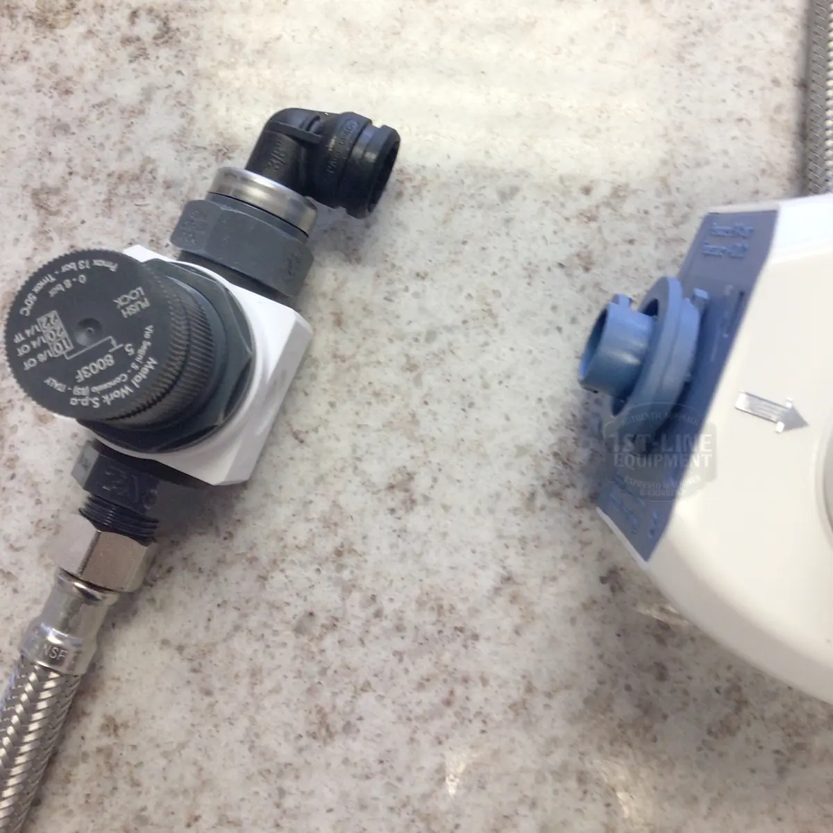 Close-up of two pressure control valves with hoses on a speckled countertop. One, from the BWT Bestmax Installation Kit, has a black dial and fittings; the other features a blue cap and white housing.