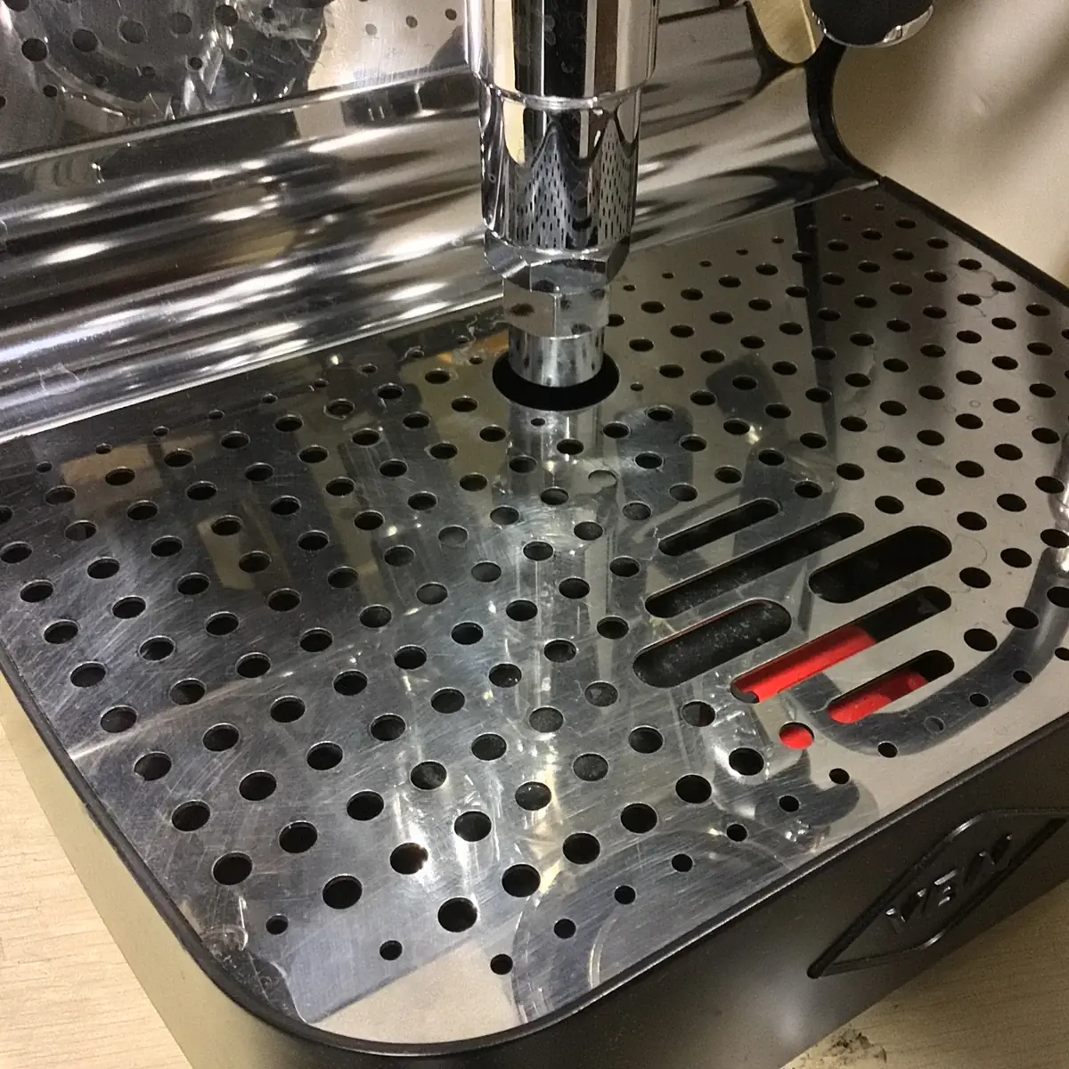 Close-up of a shiny metal drip tray with circular and elongated holes on the SIDEWALK SALE - VBM Domobar Super Analogic HX Espresso Machine OPEN BOX (E013), reflecting the machine; red indicator lights glow through the tray.