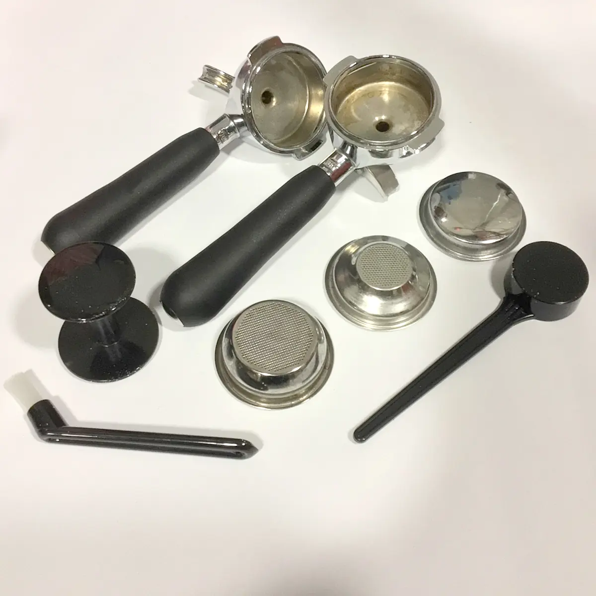 A selection of espresso machine parts—two black-handled portafilters, filter baskets, tamper, dosing tool, and spout—arranged on white surface for the SIDEWALK SALE - VBM Domobar Super Analogic HX Espresso Machine OPEN BOX (E013).