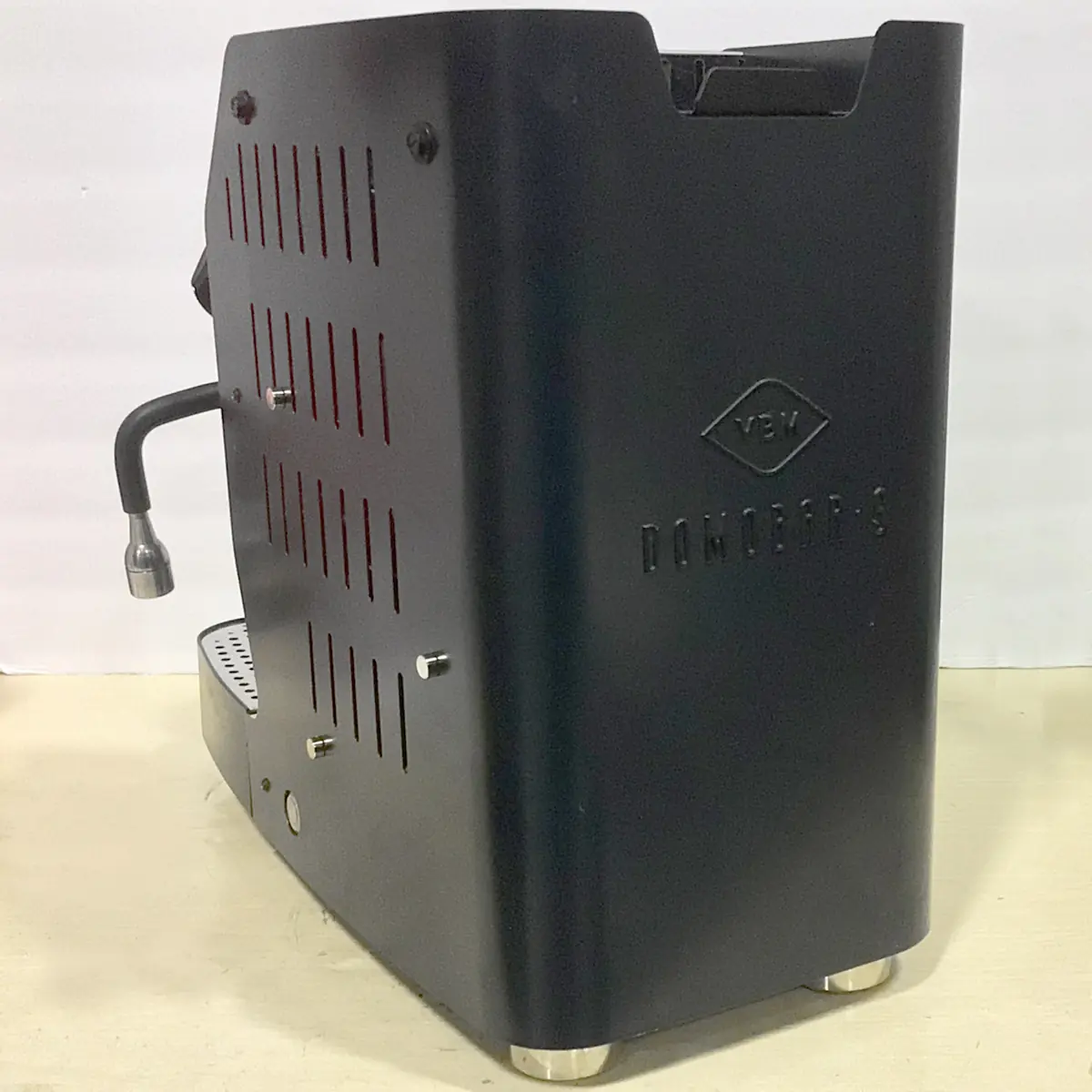Open Box SIDEWALK SALE - VBM Domobar Super Analogic HX Espresso Machine (E013) in black, shown side view with vertical vents, steam wand, metal feet, and "VEW DOMOBAR S" embossed on the panel. Machine rests on a beige surface.