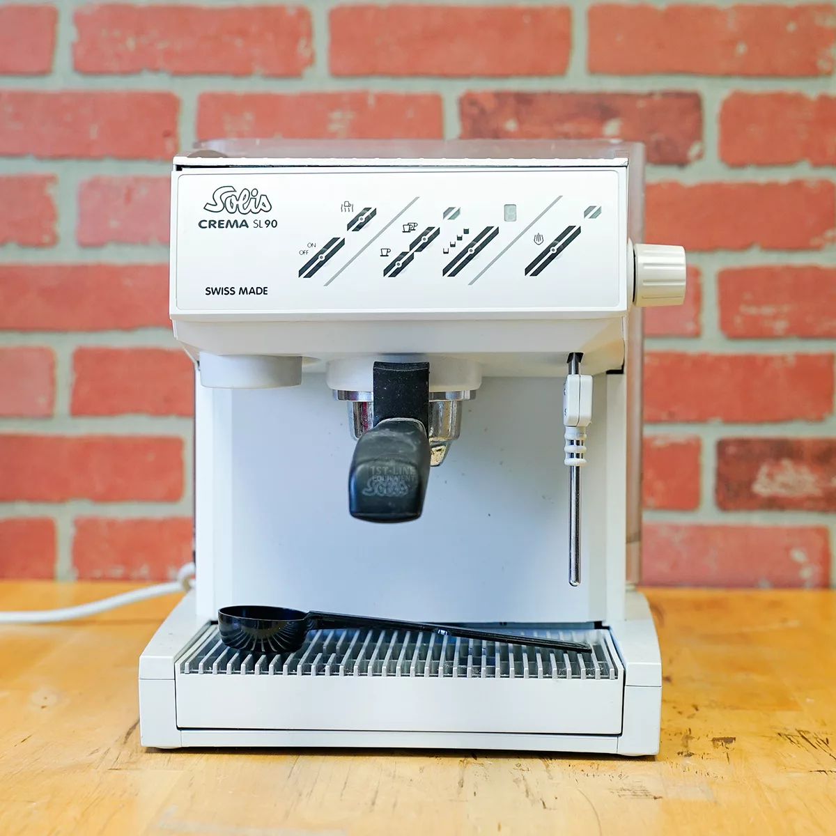 A white SIDEWALK SALE Solis Crema SL 90 (FOR PARTS ONLY) (E109) espresso machine with buttons and a steam wand sits on wood by a brick wall, a black spoon on its drip tray—great parts find!. © 2025 1st-line.com