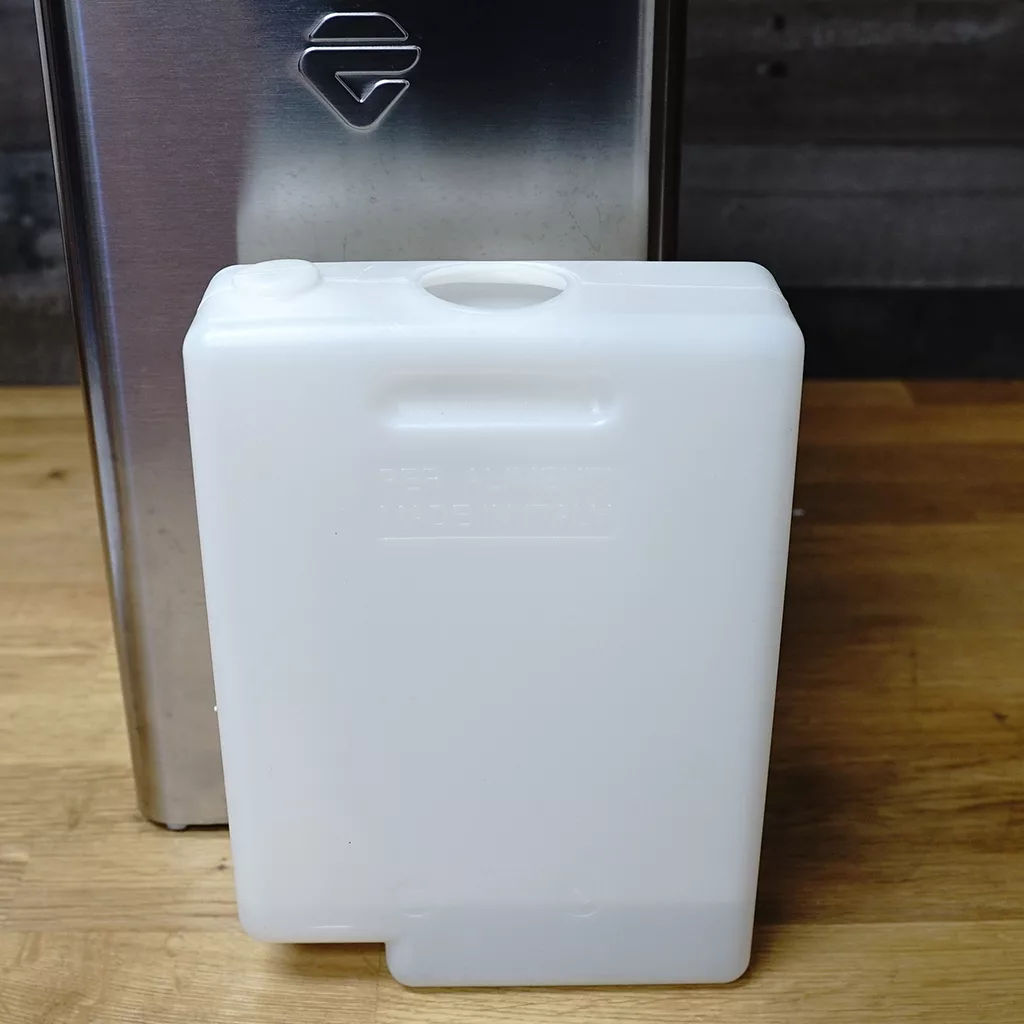 A white rectangular plastic water reservoir sits on a wooden surface in front of a stainless steel appliance, with a circular opening visible on top. © 2025 1st-line.com