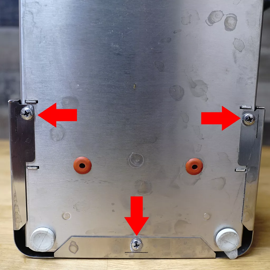 Close-up of a metal appliance back panel with three large red arrows pointing to screws—two on the upper left and right sides, and one at the center bottom—indicating areas for removal. © 2025 1st-line.com