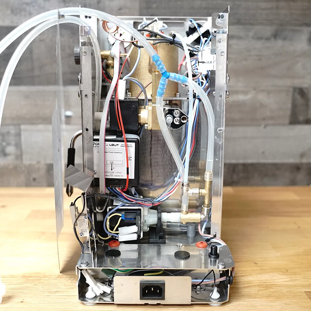 A partially disassembled espresso machine showing internal components, wiring, tubes, and metal frame, with parts exposed and visible on a wooden surface against a wooden wall background. © 2025 1st-line.com