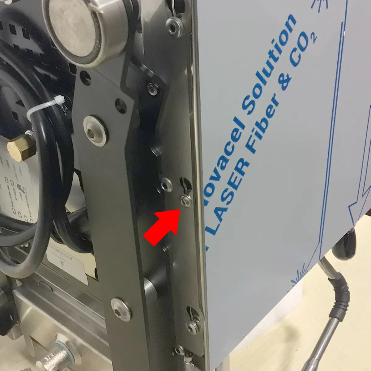 Close-up of industrial equipment, similar to the inside access of a Bezzera Sole espresso machine, with a protective blue film labeled Novacel Solution LASER Fiber & CO₂. A red arrow points to a small screw; cables and components are visible.