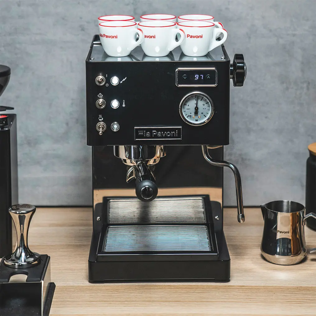 A black La Pavoni Casabar PID Espresso Machine sits on a wooden counter with three white espresso cups on top, a milk frothing pitcher and coffee tamper nearby. The machine features intuitive buttons, a gauge, and a digital display.