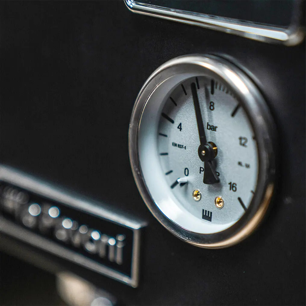 A close-up of a round pressure gauge with a white dial and black numbers on the La Pavoni Casabar PID Espresso Machine displays pressure in bars. Mounted on a black surface, the needle points slightly above 1 bar.