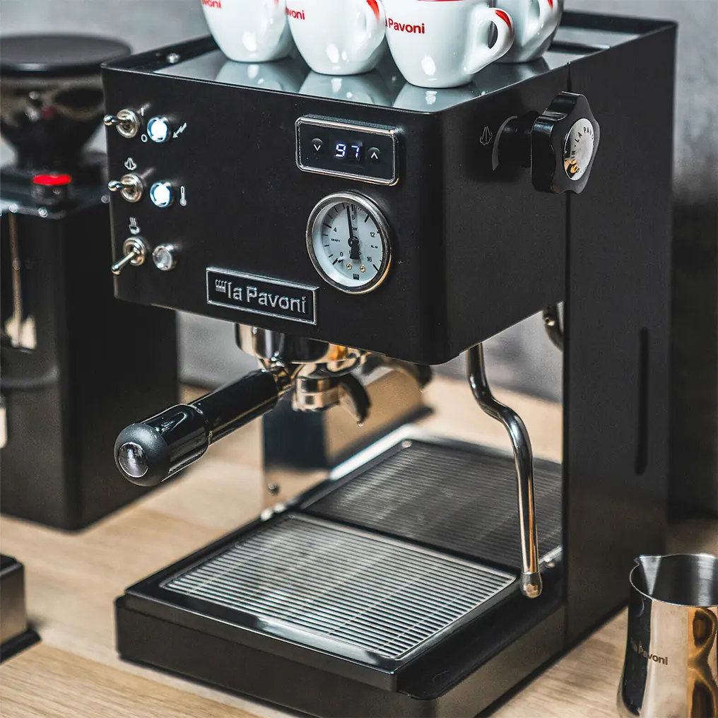 A La Pavoni Casabar PID Espresso Machine in black, featuring a pressure gauge, portafilter, and control buttons, sits on a wooden counter with several espresso cups stacked on top.