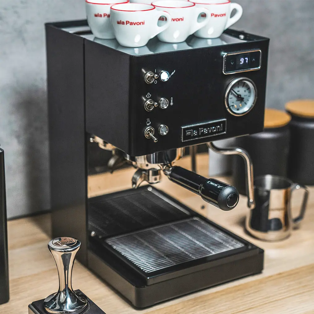A sleek black La Pavoni Casabar PID Espresso Machine sits on a wooden countertop, with three white cups on top and a metallic tamper nearby. It features buttons, a pressure gauge, and a digital temperature display.