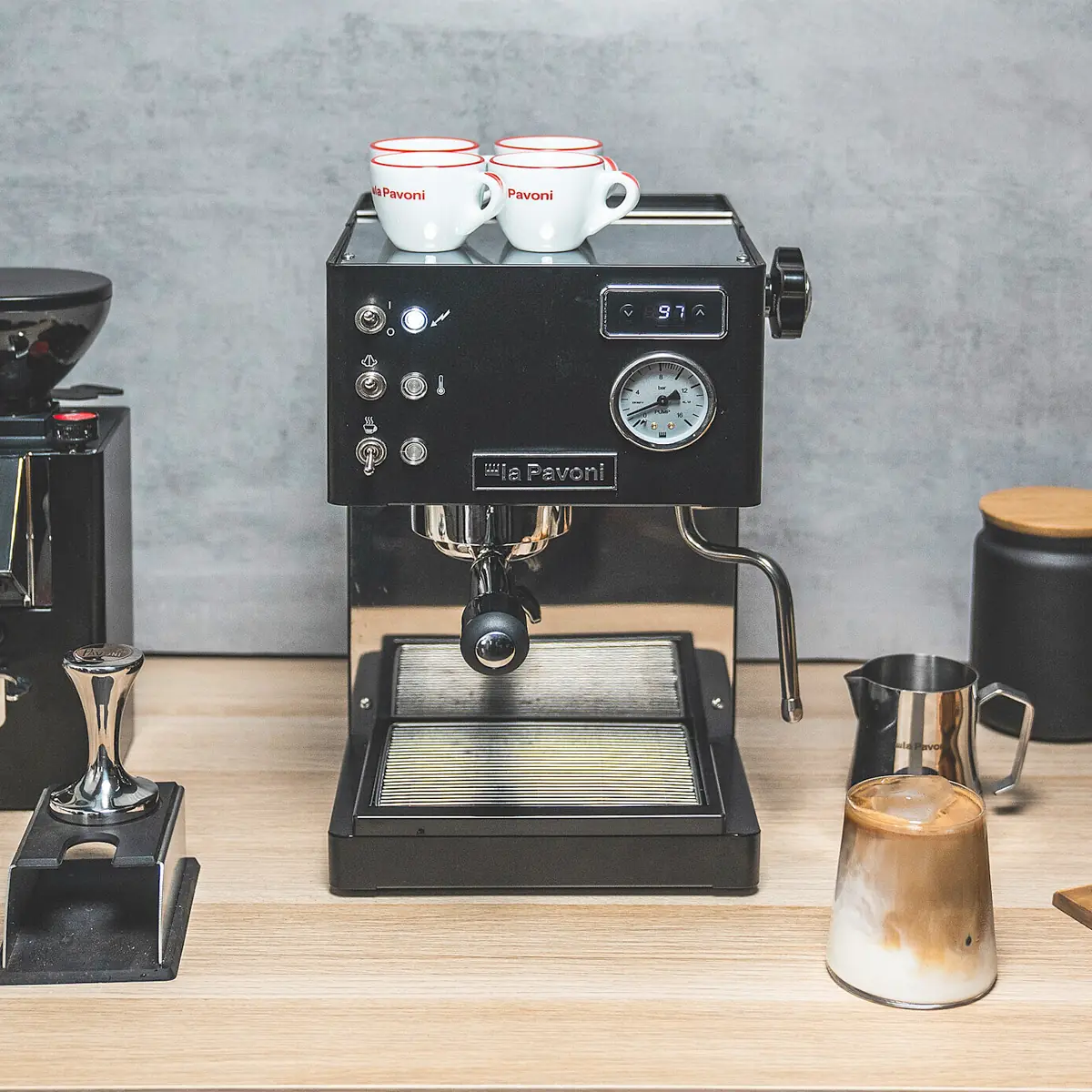 A La Pavoni Casabar PID Espresso Machine sits on a wooden countertop with two espresso cups on top, surrounded by a coffee grinder, milk frothing pitcher, tamper, and black canister.
