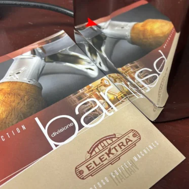 A folded mirror rests on a coffee machine brochure, reflecting and distorting the image and text, creating an illusion of misaligned letters and shapes on a wooden surface.