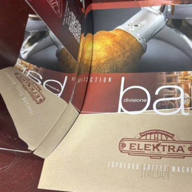 A brown and tan Elektra espresso coffee machine brochure lies open on a table, displaying a close-up image of a hand holding a portafilter. The Elektra logo is visible on the front cover.