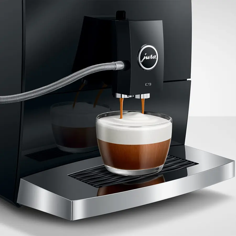 The Jura C9 Super-Automatic Espresso Machine pours espresso into a clear glass with a thick frothy milk layer, all set on its sleek metallic drip tray.