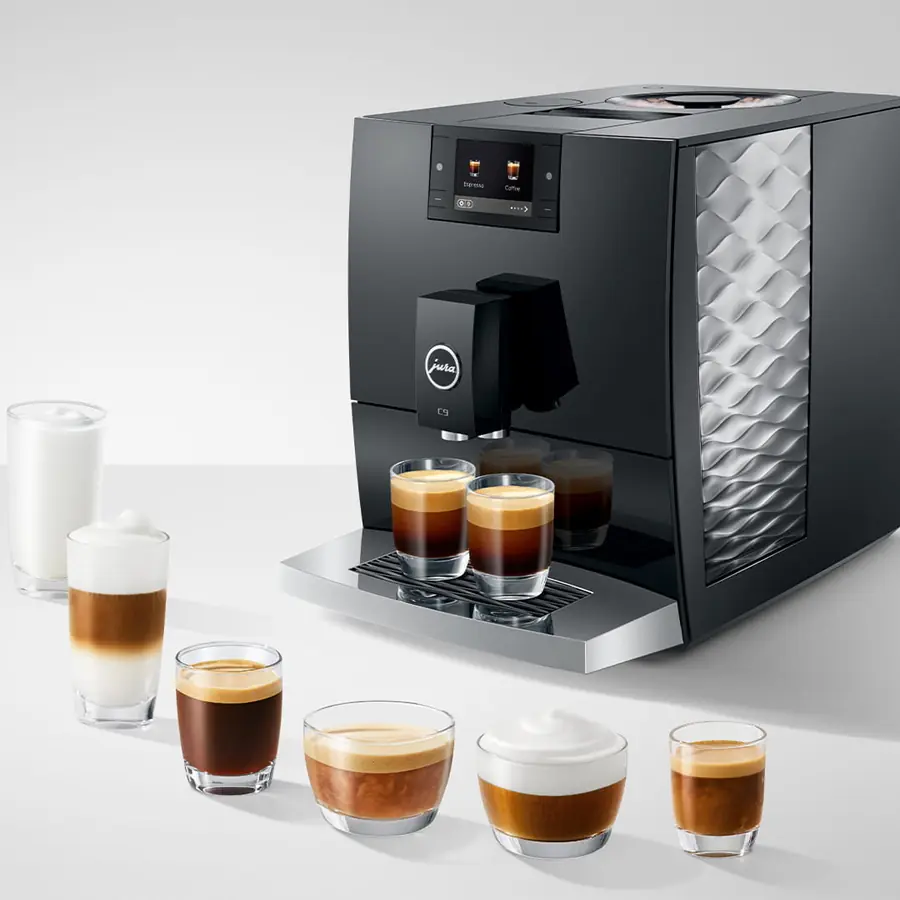The Jura C9 Super-Automatic Espresso Machine brews a variety of espresso drinks into six glass cups, each displaying distinct coffee and milk layers. A tall glass of frothed milk sits beside it on a white surface.