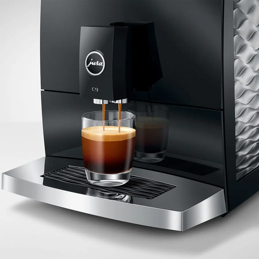 The Jura C9 Super-Automatic Espresso Machine, in sleek black and silver, pours espresso into a clear glass, highlighting creamy coffee with a rich crema layer on top.