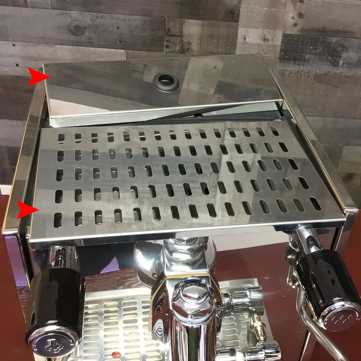 A close-up of a shiny Magister Stella espresso machine with two black handles, a slotted metal drip tray, and two red arrows pointing to a gap between the drip tray and the machine's frame. Inside, a wooden wall serves as the background.