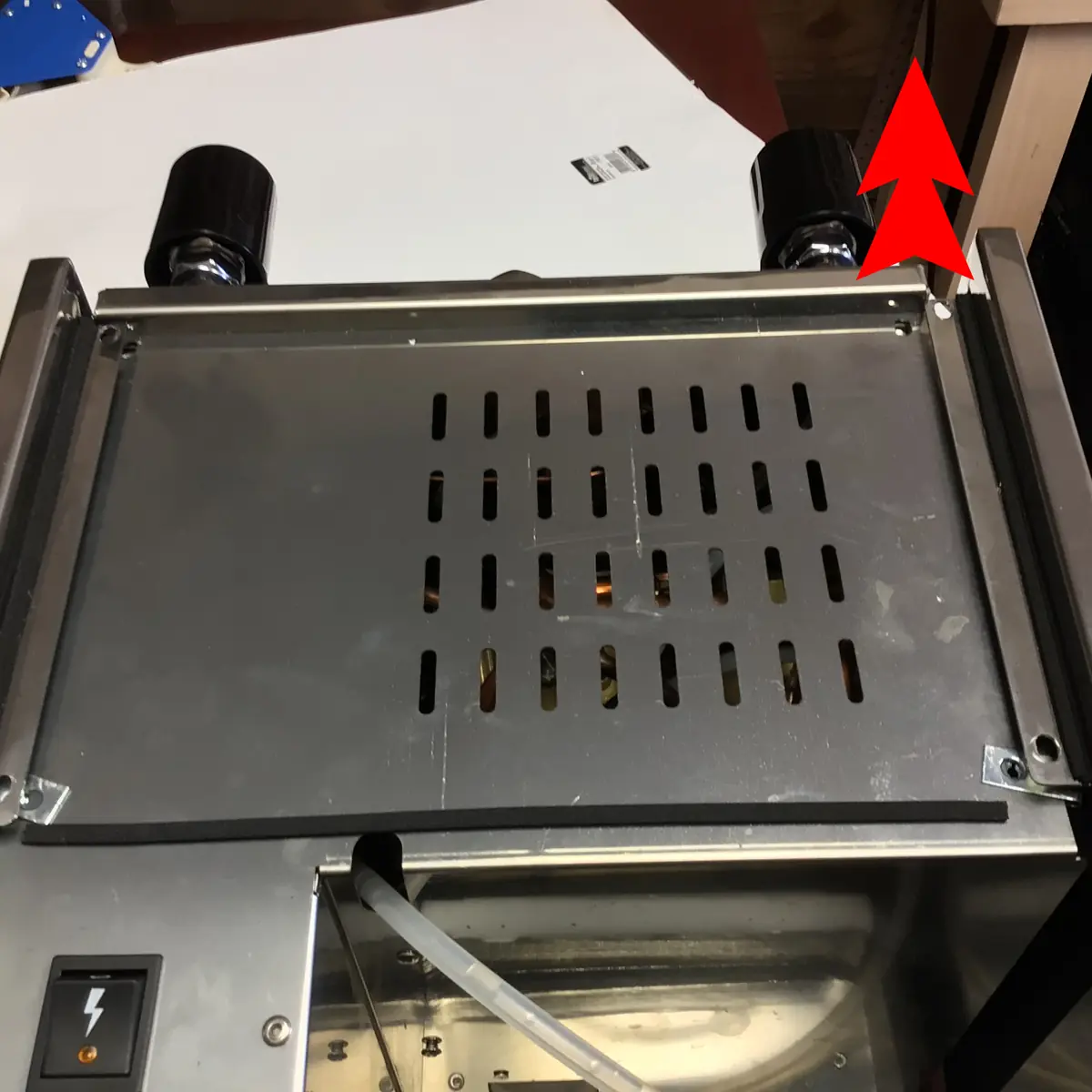 A Magister Stella metal machine with a grated surface and two black knobs on top. A switch and a cable are visible at the bottom left, with a large red arrow pointing to Accessing the Inside in the upper right corner.