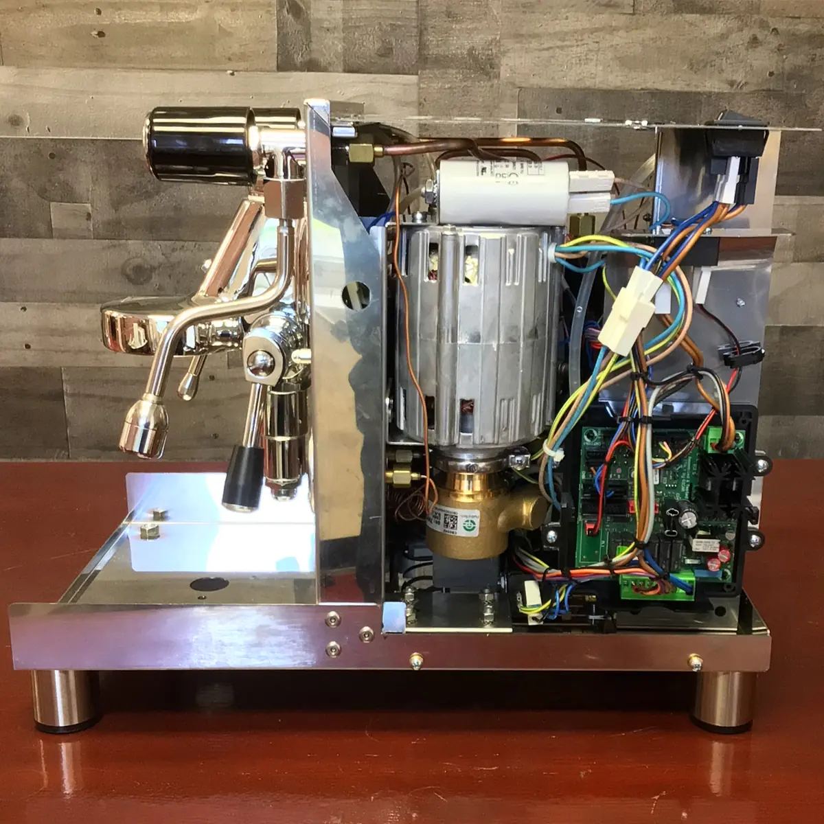Accessing the inside of a Magister Stella semi-automatic espresso machine with its right side panel removed exposes internal components like wiring, circuit boards, a motor, and metal pipes, all set on a wooden surface with a wood-patterned wall background.
