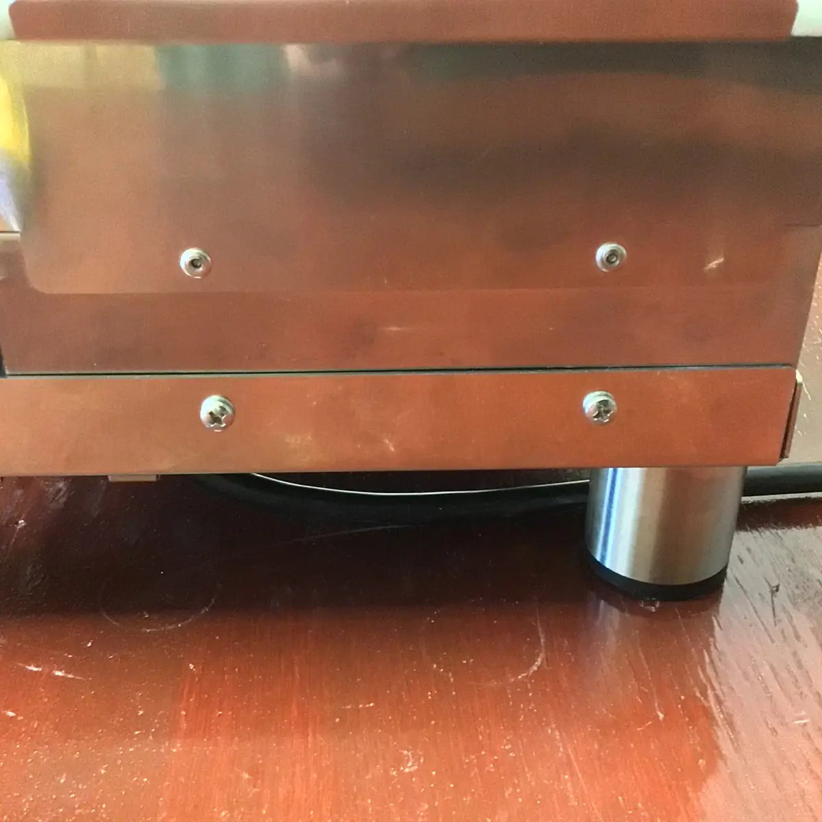 A close-up of a Magister Stella stainless steel appliance with visible screws and a cylindrical metal leg, placed on a shiny wooden surface. A black power cord is partially visible behind the appliance, hinting at its inner workings inside.