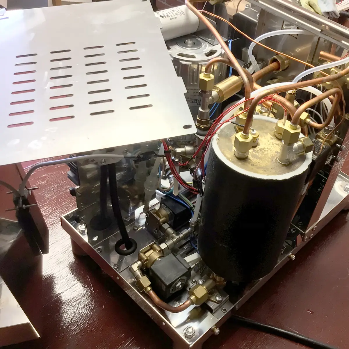 The inside of a Magister Stella espresso machine reveals copper pipes, a cylindrical boiler, brass fittings, and wiring, all visible with the metal cover partially removed on a brown tabletop.