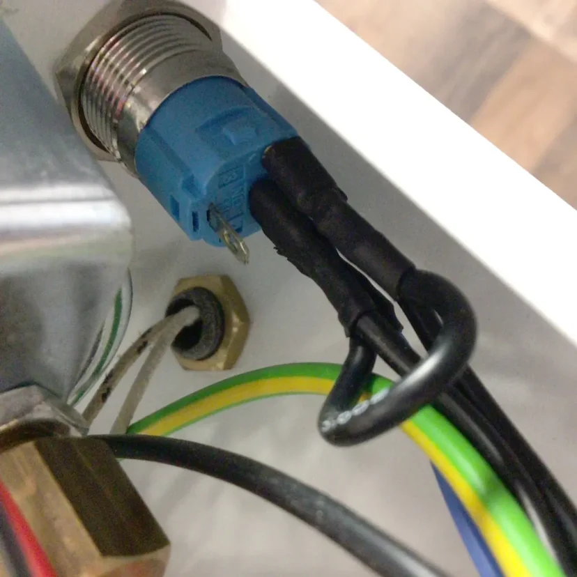Close-up of a blue electrical connector with black wires attached inside a Nurri R-Type enclosure. Yellow-green and white wires are also visible, along with various metal components near the power button of the dual boiler system.
