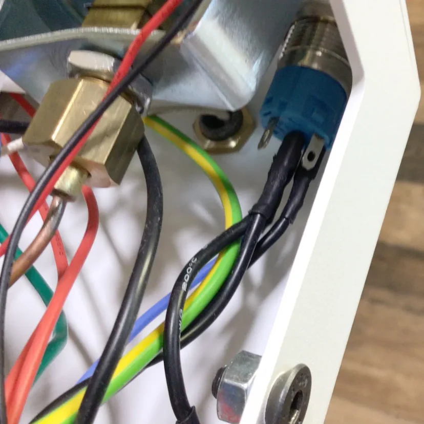 Close-up of electrical wires (red, yellow, green, black) connected to terminals inside a white metal enclosure of a Nurri R-Type Dual Boiler machine. A blue component and brass fitting are visible; background features a wooden surface.
