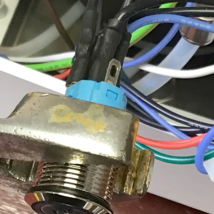 Close-up of a metallic component with a threaded end, attached to colorful wires—including blue, green, red, and black—connected by a blue plastic connector. In the background, more wires and part of a Nurri R-Type dual boiler device are visible.