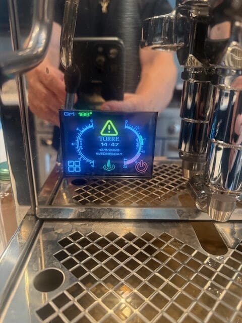 Close-up of a digital display on a coffee machine showing a warning triangle, the word TORRE, temperature, date, and other indicators, with a persons reflection partially visible in the background.