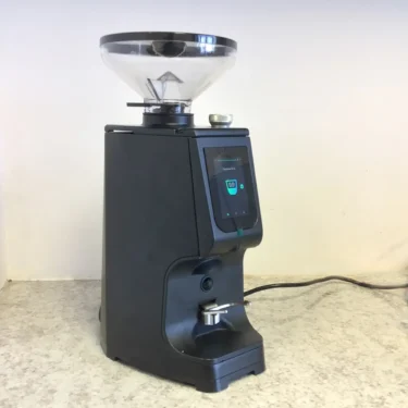 A SIDEWALK SALE LUCCA Atom 75 Espresso Coffee Grinder in black, featuring a clear bean hopper and digital display, sits on a speckled countertop with its power cord plugged in at the back.