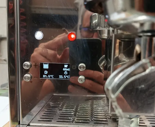 A person takes a photo of a coffee machine with metal surfaces, showing a digital display indicating 40 ml, 81.6°C, and 32.5°C, along with illuminated buttons and a reflection of the person holding a phone.
