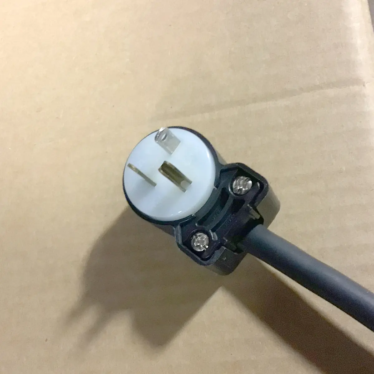 A close-up of a three-prong black electrical plug, set against plain cardboard—an essential accessory for your SOLD SIDEWALK SALE - Orchestrale Nota White HX Espresso Machine (E111), ensuring safe power at any event.