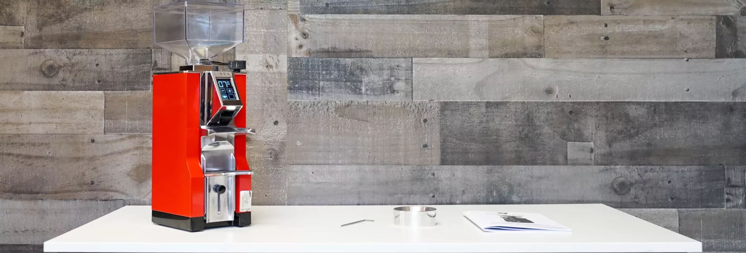 A bright red Eureka Mignon Libra Espresso Coffee Grinder - Weight-Based Grinding sits on a white table by a rustic wood wall, alongside a metal ring, a small tool, and an open instruction manual.