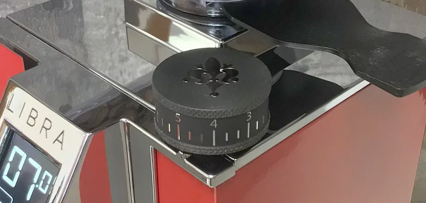 Close-up of a Eureka Mignon Libra Espresso Coffee Grinder - Weight-Based Grinding in metallic red, showing the black fleur-de-lis adjustment dial set between 3 and 4, plus part of the “LIBRA” weight-based grinding display.