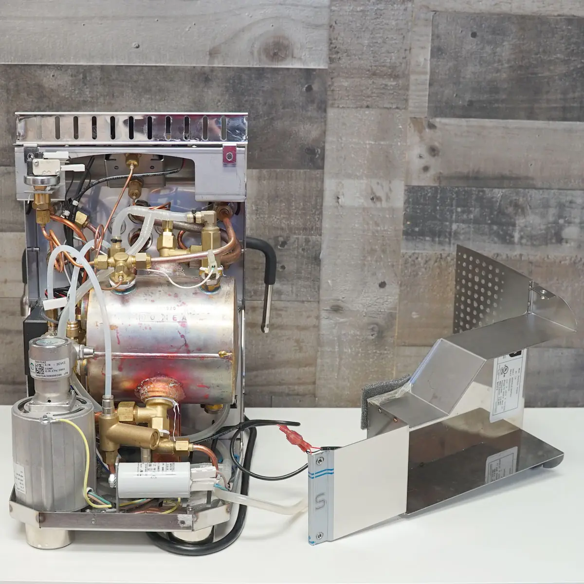 A VBM Domobar espresso machine with its metal casing removed, revealing complex internal components like wires, tubes, valves, and a boiler. The Super Analogic HX metal side panel rests beside the exposed machine on a white surface.