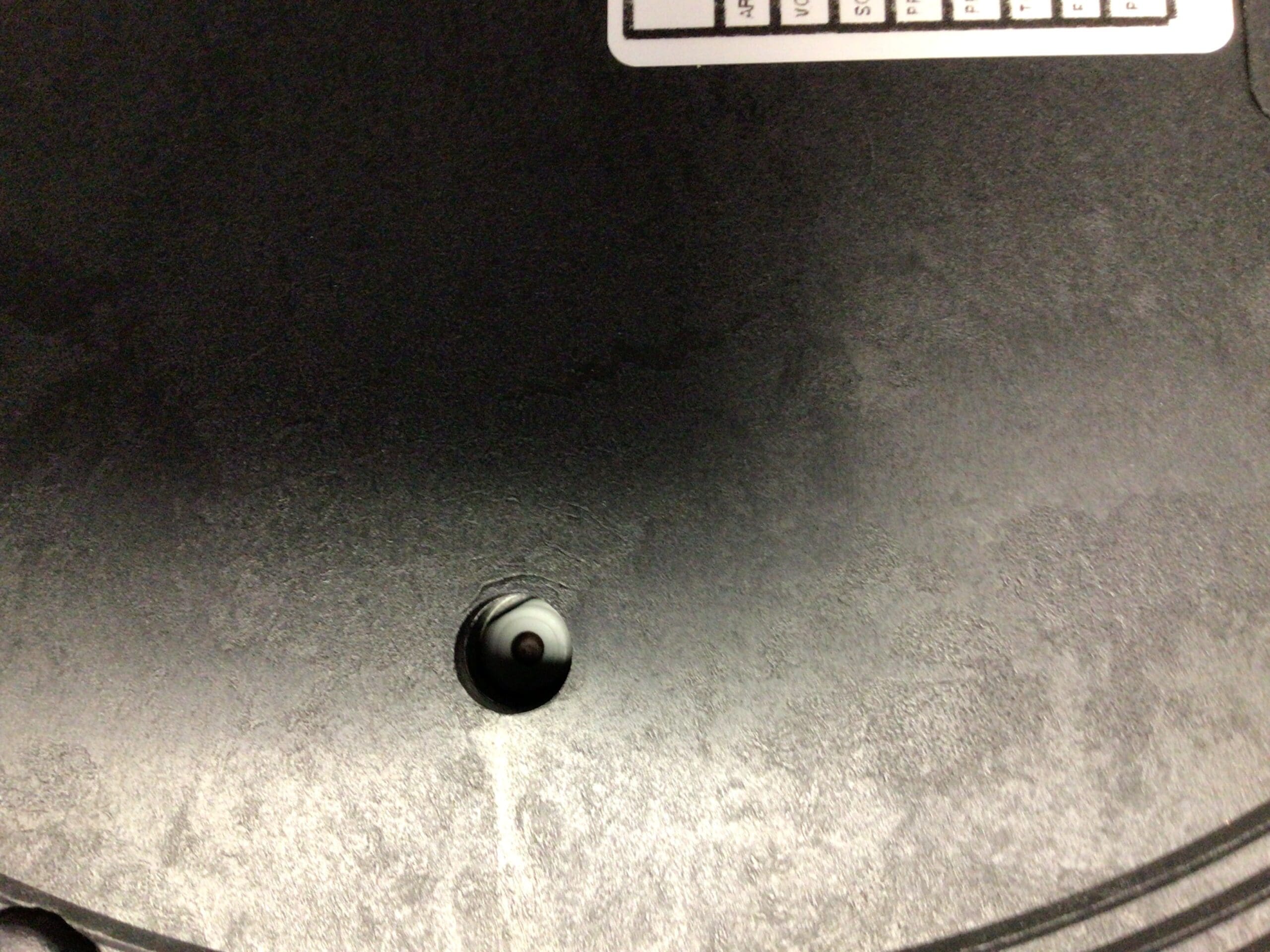 Close-up of a black surface with a small round hole near the bottom center, revealing a white and black object—possibly part of an Elektra Microcasa a Leva Safety Thermostat Reset. A white sticker with a grid and text is visible at the images top edge. © 2025 1st-line.com