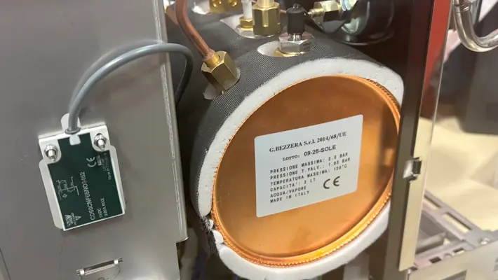 A close-up of a cylindrical metal component from the Bezzera Sole PID HX Espresso Machine with a label showing technical details. Wires and tubes are connected, and a green circuit board is visible nearby.