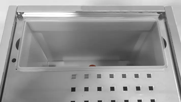 A close-up of an empty rectangular stainless steel container with a clear plastic lid, set in a countertop with a perforated metal surface—perfect for use with your Bezzera Sole PID HX Espresso Machine.