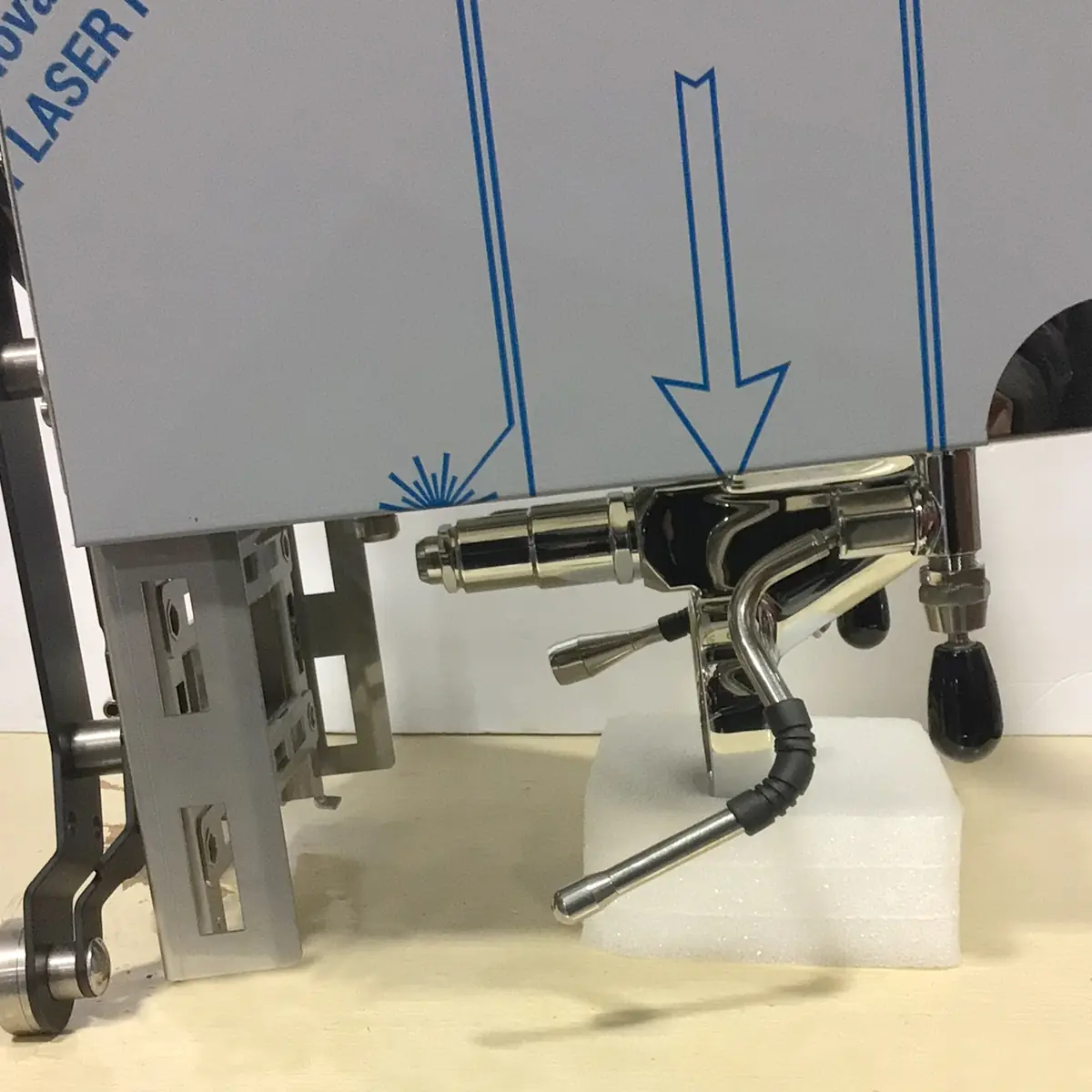 A close-up of a shiny Bezzera Sole espresso machine group head and steam wand, partially wrapped in protective film with blue arrows, resting on a white foam block on a light surface.