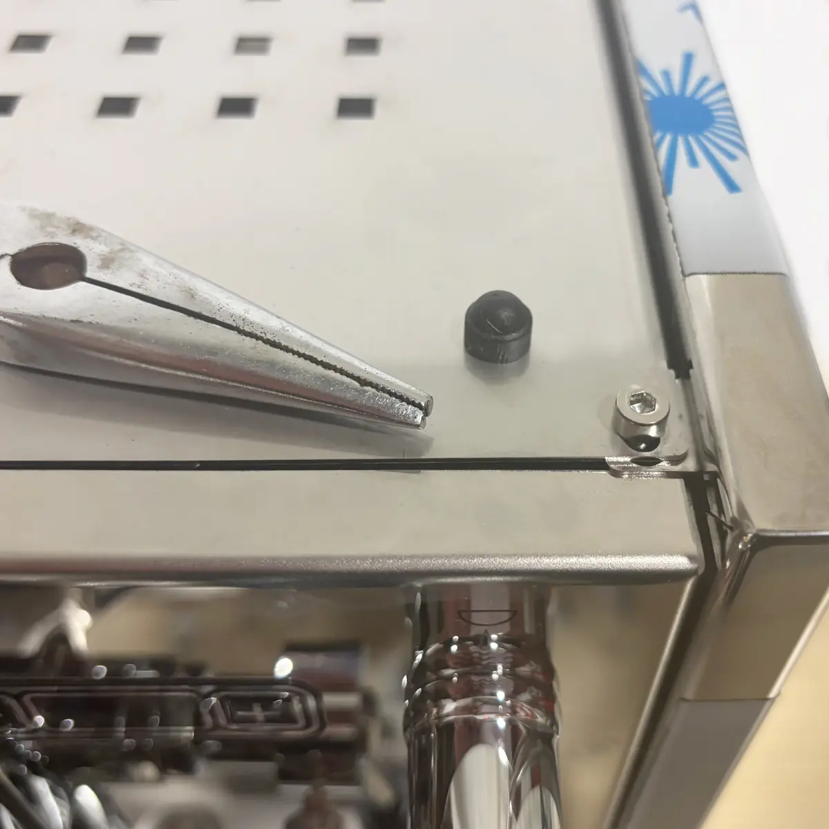 A pair of needle-nose pliers rests on a stainless steel espresso machine, near a small black rubber cap and a metal screw. The edge of the Bezzera Sole and a blue marking hint at inside access for maintenance or repair.