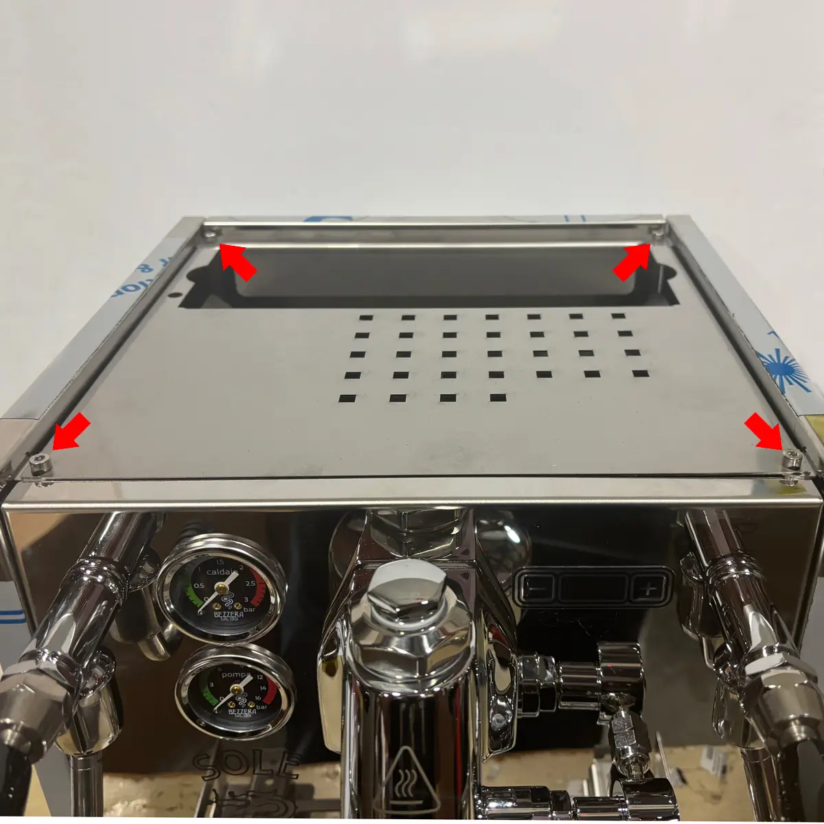 A close-up of a stainless steel Bezzera Sole espresso machine shows four red arrows pointing to each corner of the top removable panel, indicating points for accessing inside. Two pressure gauges and control knobs are visible on the front.