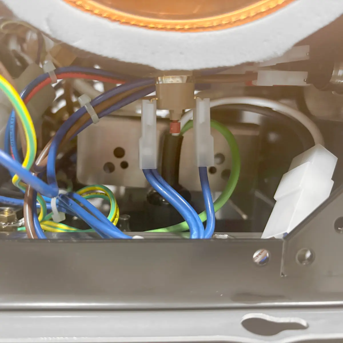 Close-up of electrical wiring inside a metallic appliance, showing blue, green-yellow, and brown wires connected to white plastic connectors, with mechanical components partially visible in the background.