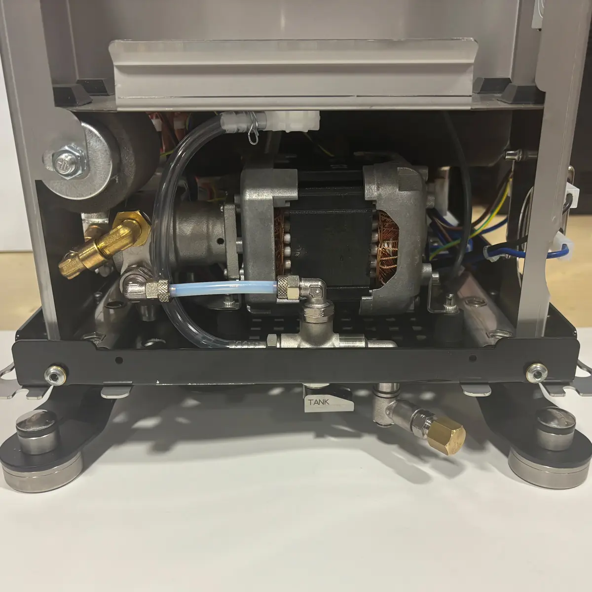 The image shows the internal components of a machine, featuring a motor, wires, tubes, and metal fittings. The machine is elevated on four legs, and a label reading TANK is visible near the bottom center.