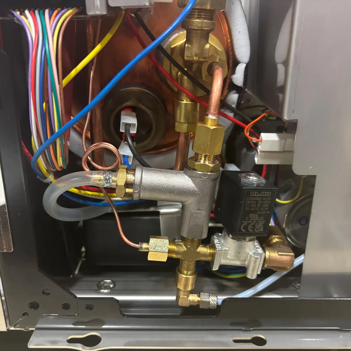 Close-up view of the internal components of a machine, showing colorful wires, copper tubing, brass fittings, a solenoid valve, and various connectors arranged within a metallic enclosure.