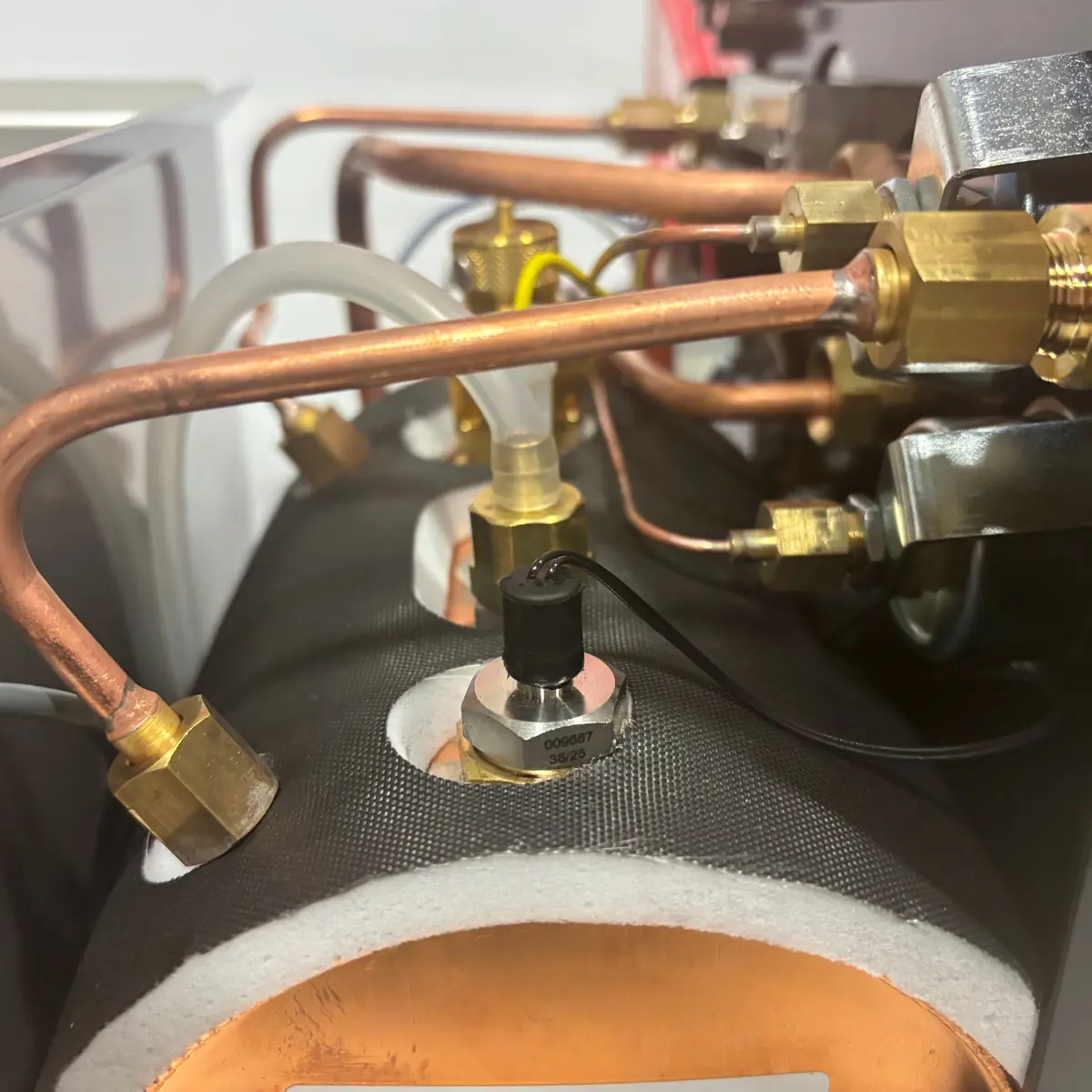 Close-up of copper pipes, brass fittings, and a black sensor attached to a cylindrical, insulated metal component, possibly part of industrial or HVAC equipment. White and clear tubes are also connected.