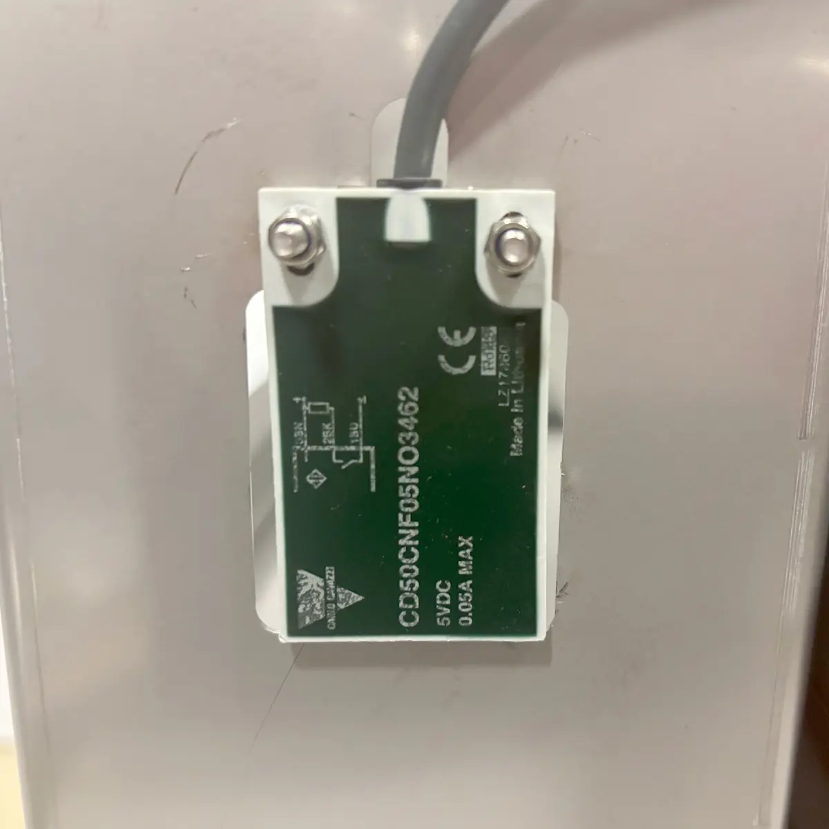 Close-up of a green electronic sensor or module labeled CDS5CNFE0503462, attached to a white surface with two screws. A black cable is connected at the top, and technical specifications are visible.