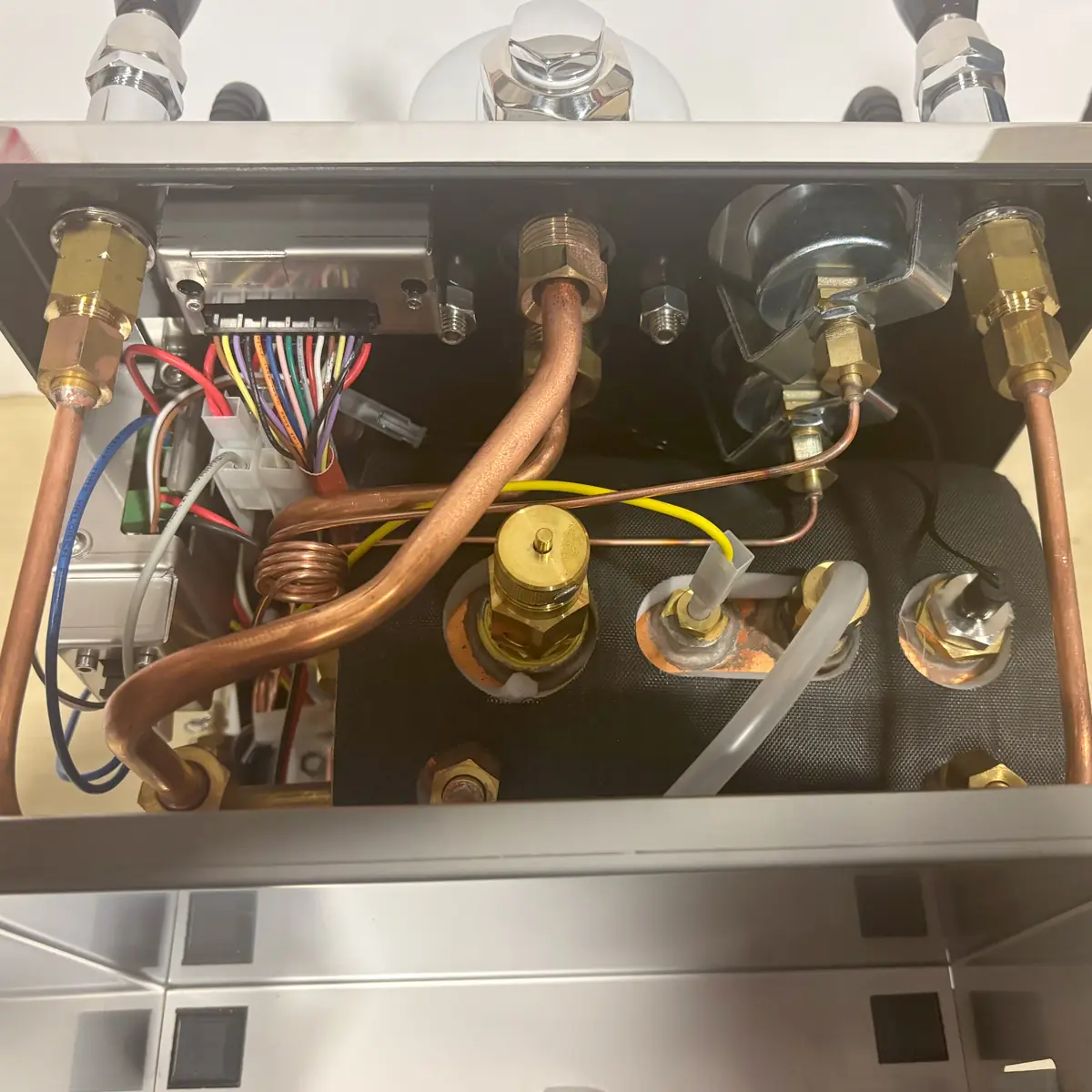 Close-up view of the internal components of a machine, showing copper pipes, brass fittings, wires, and tubes connected to a black insulated part and circuit board, likely part of an espresso machine or similar appliance.