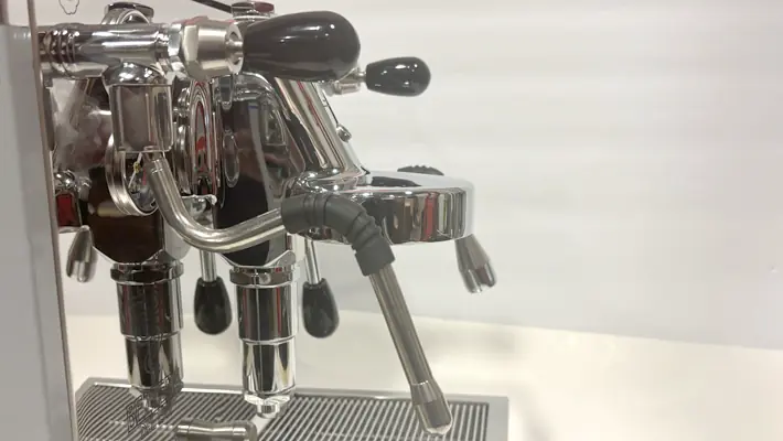 A close-up view of a shiny espresso machine with multiple levers and spouts, positioned on a metal drip tray against a plain white background.