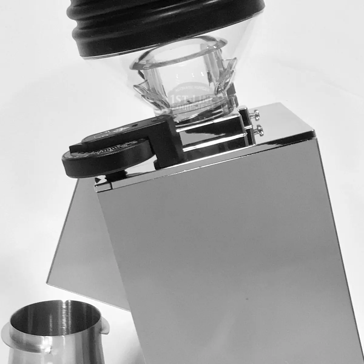 A black and white image of the Eureka Mignon Single Dose Pro Espresso Coffee Grinder with a glass hopper on top. A small metal cup is placed beside the grinder on a white surface.