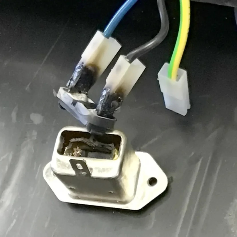 A close-up of a damaged electrical connector with melted and blackened plastic, attached to three wires—blue, black, and yellow with a green stripe—on a dark surface.
