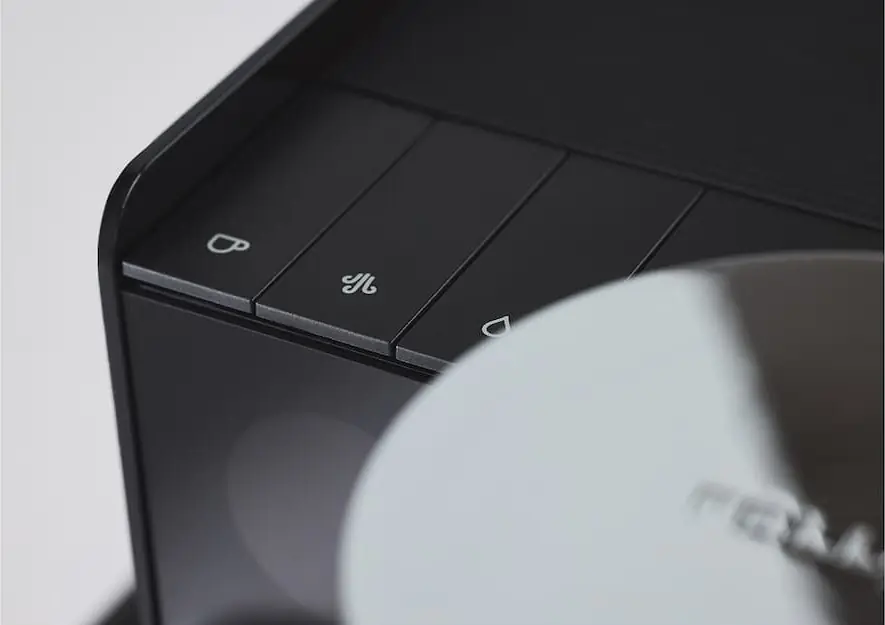 Close-up of three black rectangular buttons on the Fellow Series 1 Espresso Machine, each with a white icon: a cup, a steam symbol, and a droplet. A blurred silver circular object appears in the foreground.
